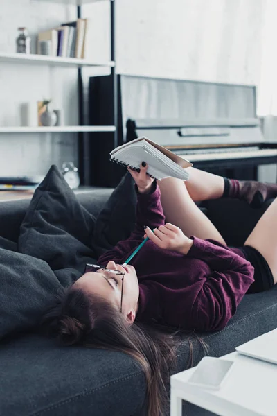 Schönes Mädchen mit Brille auf Sofa liegend, Bleistift in der Hand und Blick auf Notizbuch im Wohnzimmer — Stockfoto