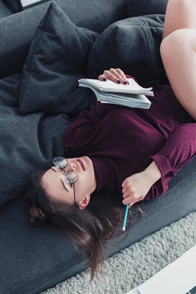 Attraktives Mädchen, das auf der Couch liegt und Notizbuch und Bleistift in der Hand hält — Stockfoto