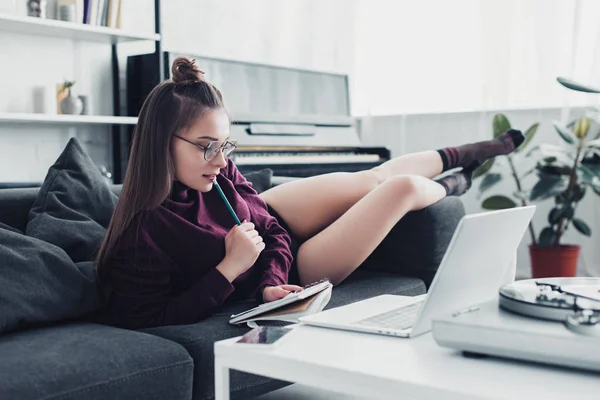 Fille réfléchie couchée sur le canapé et tenant un cahier et un crayon à la maison — Photo de stock