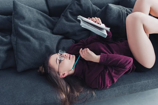 Chica atractiva acostada en el sofá y la celebración de cuaderno y lápiz en la sala de estar - foto de stock