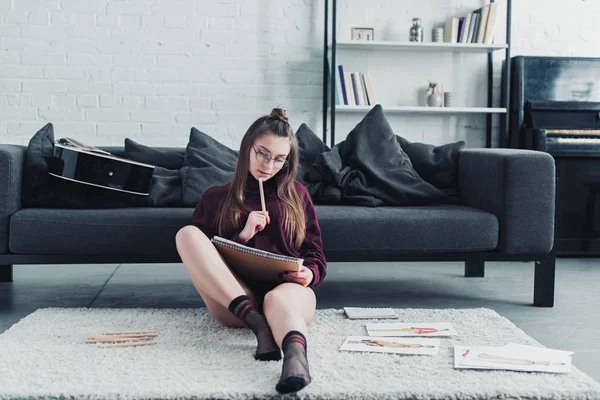 Attraktive Designerin sitzt auf Teppich neben Sofa und hält Bleistift und Notizbuch im Wohnzimmer — Stockfoto