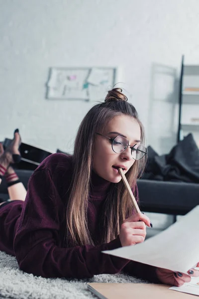 Beautiful designer lying on carpet, biting pencil and looking at sketch in living room — Stock Photo