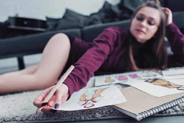 Beautiful designer lying on carpet and holding pencil and sketch at home — Stock Photo