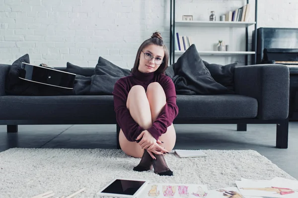 Hermoso diseñador sentado en la alfombra cerca del sofá y mirando a la cámara en la sala de estar — Stock Photo