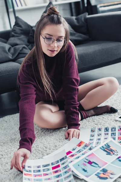 Schöne Fotografin in Brille sitzt auf Teppich neben Couch und betrachtet Fotos im Wohnzimmer — Stockfoto