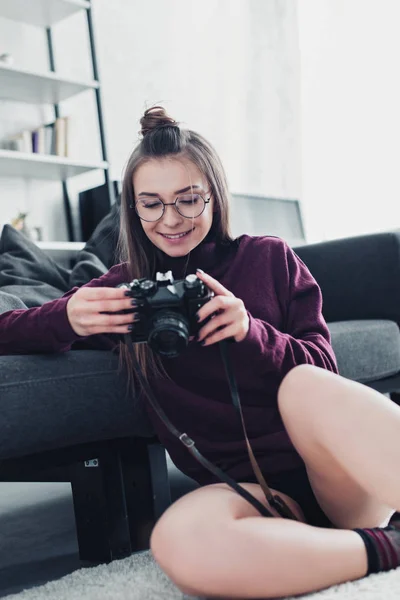 Привлекательный фотограф улыбается, сидит на полу рядом с диваном и держит пленочную камеру в гостиной — стоковое фото