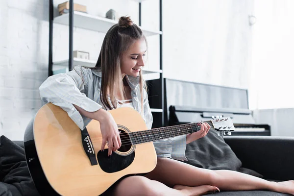 Hermoso músico sentado en el sofá, sonriendo y tocando la guitarra acústica en casa - foto de stock