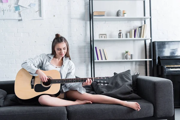 Beautiful musician sitting on sofa and playing acoustic guitar at home — Stock Photo