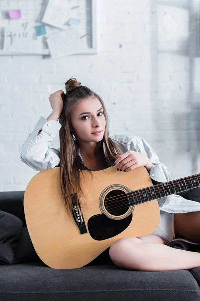 Attraktive Musikerin sitzt auf Sofa, hält Akustikgitarre in der Hand und schaut zu Hause in die Kamera — Stockfoto