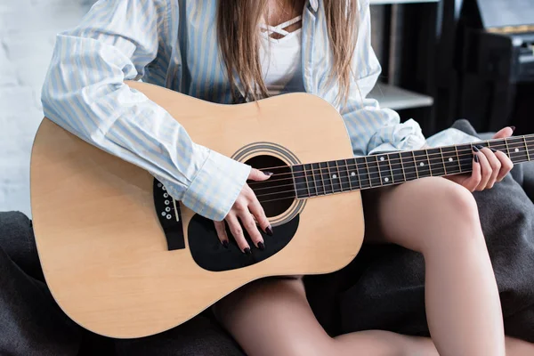 Vista cortada do músico sentado no sofá e tocando guitarra acústica em casa — Fotografia de Stock