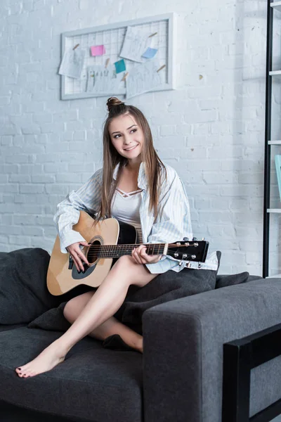 Musicista sorridente seduto sul divano e suonare la chitarra acustica a casa — Foto stock