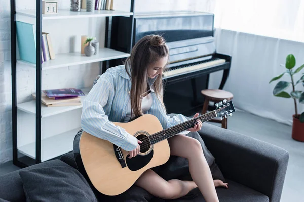 Музыкант сидит на диване и играет на акустической гитаре в гостиной — стоковое фото