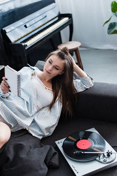 Attraktives Mädchen, das auf dem Sofa in der Nähe eines alten Plattenspielers liegt und zu Hause Notizbuch liest — Stockfoto