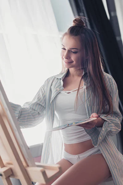 Sexy Künstlerin in Hemd und Unterwäsche mit Palette und Blick auf Staffelei im Wohnzimmer — Stockfoto
