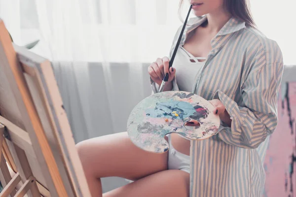 Cropped view of artist holding palette, biting paintbrush and sitting in front of easel in living room — Stock Photo