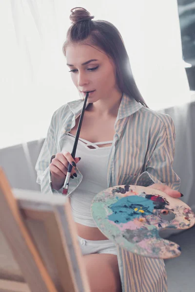 Attractive artist holding palette, biting paintbrush and looking at easel in living room — Stock Photo