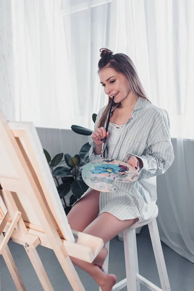 Attraktive Künstlerin sitzt auf Stuhl, hält Palette, beißt Pinsel und schaut Staffelei im Wohnzimmer — Stockfoto