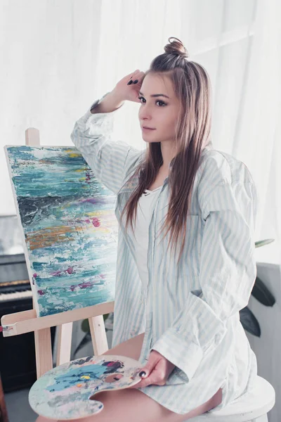 Thoughtful artist sitting on chair in front of easel and holding palette at home — Stock Photo
