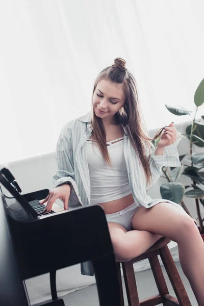Hermosa músico en camisa y ropa interior tocando el piano y sonriendo en la sala de estar - foto de stock
