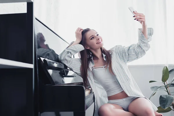 Menina bonita em camisa e roupa interior sentado no piano e tomando selfie em casa — Fotografia de Stock