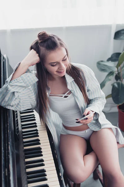 Chica sonriente en camisa y ropa interior sentado en el piano y el uso de teléfono inteligente en casa - foto de stock