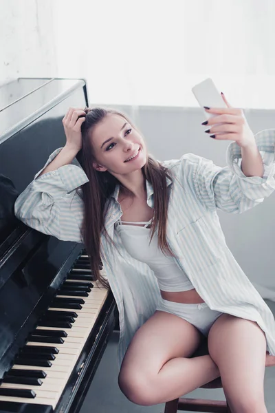 Привлекательная девушка в рубашке и нижнем белье сидит за фортепиано и делает селфи в гостиной — стоковое фото