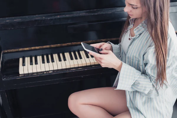 Menina atraente na camisa sentado na frente do piano e usando smartphone na sala de estar — Fotografia de Stock