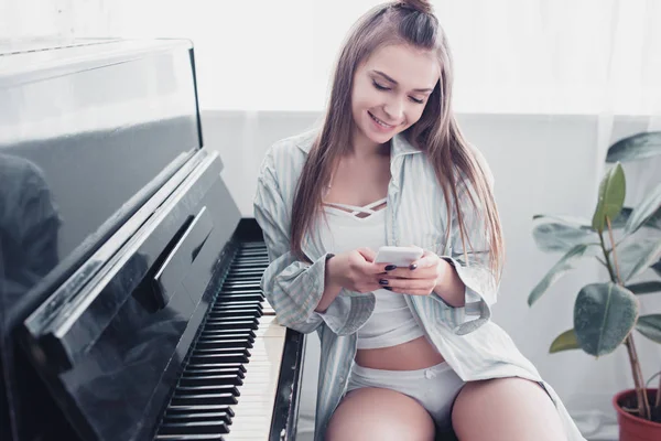 Schönes Mädchen in Unterwäsche und Hemd, das vor dem Klavier sitzt und das Smartphone im Wohnzimmer benutzt — Stockfoto