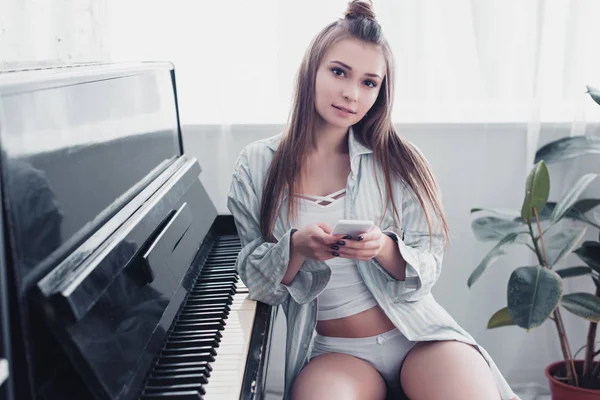 Приваблива дівчина в нижній білизні і сорочці, сидячи перед піаніно, тримає смартфон і дивиться на камеру у вітальні — стокове фото