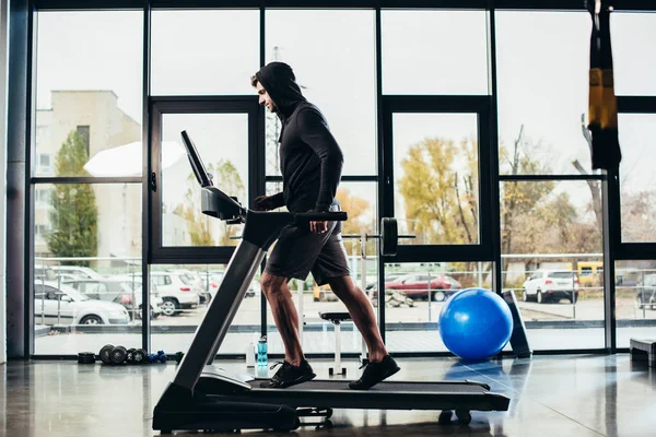 Vista lateral de deportista guapo en sudadera con capucha ejercicio en la cinta de correr en el gimnasio - foto de stock