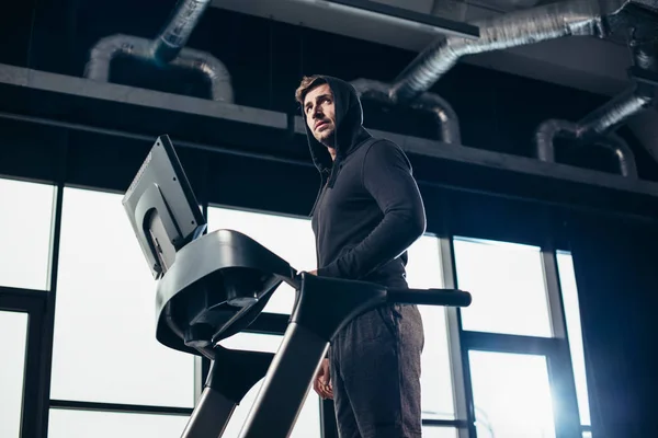 Low angle view of handsome sportsman in hoodie exercising on treadmill in gym — Stock Photo