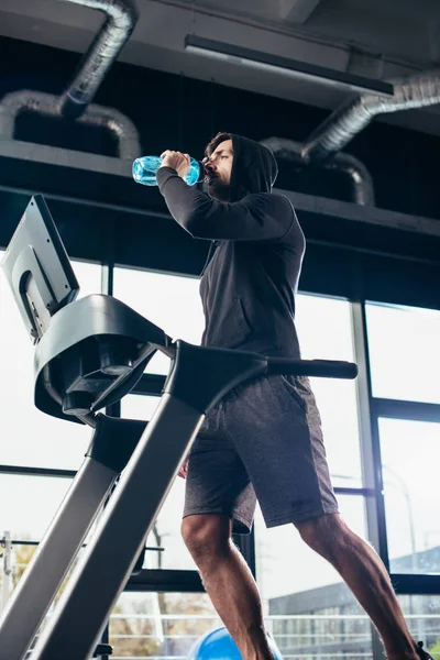 Low angle view of handsome sportsman in hoodie exercising on treadmill and drinking water in gym — Stock Photo