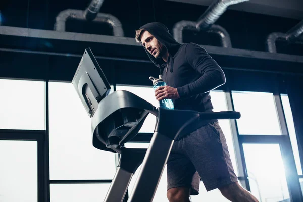 Low angle view of handsome sportsman in hoodie exercising on treadmill and holding sport bottle with water in gym — Stock Photo