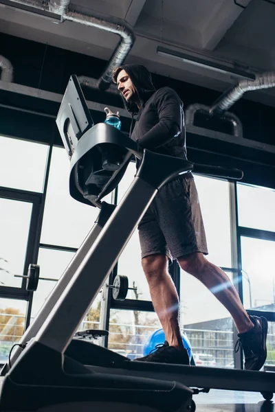 Low angle view of handsome sportsman in hoodie running on treadmill and holding sport bottle with water in gym — Stock Photo