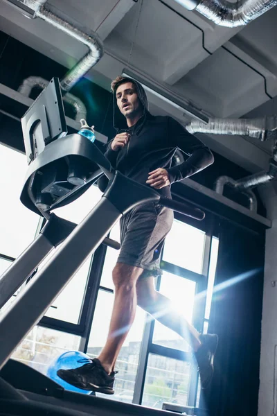 Low angle view of handsome sportsman in hoodie exercising on treadmill in gym and looking at camera — Stock Photo