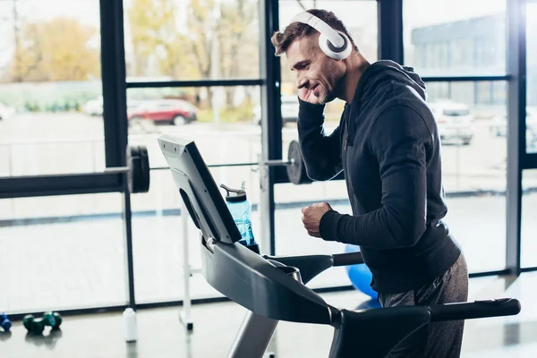 Vista lateral de deportista guapo en sudadera con capucha ejercicio en la cinta de correr y escuchar música en el gimnasio - foto de stock