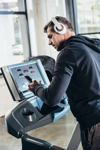 Apuesto deportista en sudadera con capucha ejercicio en la cinta de correr y el uso de teléfonos inteligentes en el gimnasio - foto de stock