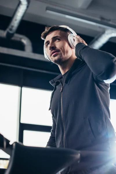 Vista de ángulo bajo de deportista guapo en sudadera con capucha ejercicio en la cinta de correr y escuchar música en el gimnasio - foto de stock