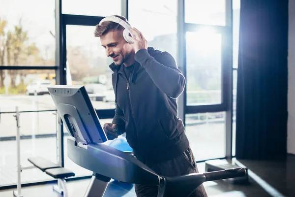 Красивый спортсмен в толстовке, упражняющийся на беговой дорожке и слушающий музыку в тренажерном зале — стоковое фото