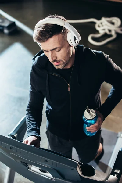 High angle view of handsome sportsman in hoodie exercising on treadmill and holding sport bottle in gym — Stock Photo