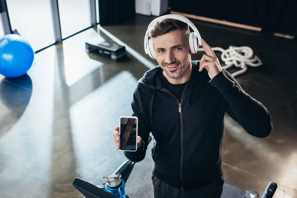 Vista de ángulo alto de sonriente deportista guapo en sudadera con capucha mostrando teléfono inteligente con pantalla en blanco en la cinta de correr en el gimnasio - foto de stock