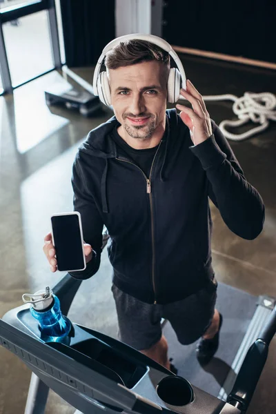 Vista de ángulo alto de deportista guapo en sudadera con capucha mostrando teléfono inteligente con pantalla en blanco en la cinta de correr en el gimnasio - foto de stock