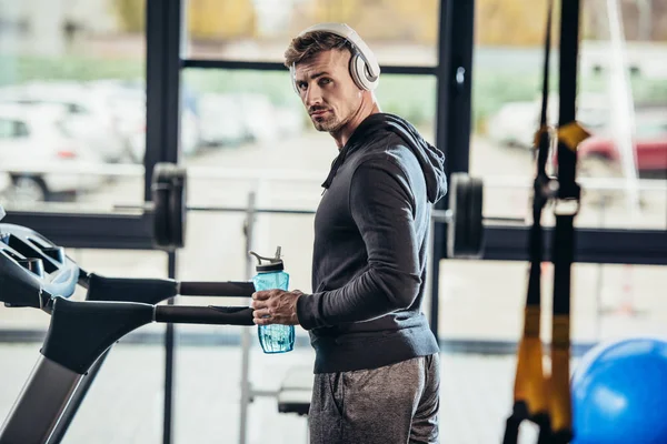 Ritratto di un bellissimo sportivo in felpa con cappuccio in piedi vicino al tapis roulant con bottiglia sportiva in palestra e guardando la fotocamera — Foto stock