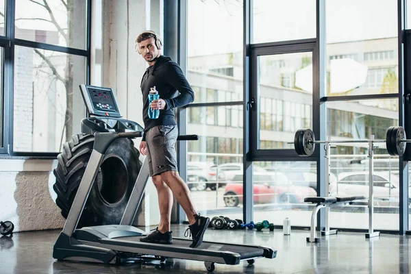 Handsome sportsman in hoodie exercising on treadmill and holding sport bottle of water in gym — Stock Photo