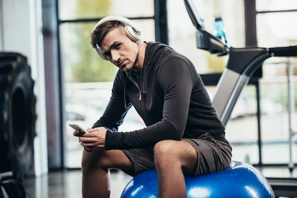 Schöner Sportler sitzt auf Fitnessball, hält Smartphone in der Hand und blickt in die Kamera im Fitnessstudio — Stockfoto