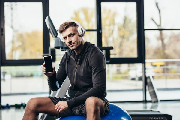 Handsome sportsman sitting on fitness ball and showing smartphone with blank screen in gym — Stock Photo