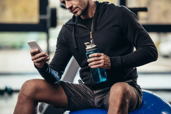 Abgeschnittenes Bild eines Sportlers, der auf einem Fitnessball sitzt und das Smartphone im Fitnessstudio benutzt — Stockfoto