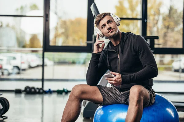 Apuesto deportista sentado en la pelota de fitness y escuchar música con smartphone en el gimnasio - foto de stock