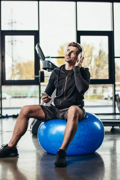 Sportif beau et joyeux assis sur le ballon de fitness et écouter de la musique avec smartphone dans la salle de gym — Photo de stock