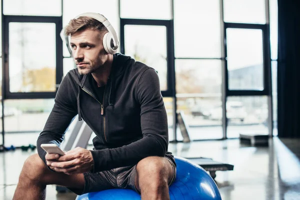 Apuesto deportista sentado en la pelota de fitness con teléfono inteligente en el gimnasio - foto de stock